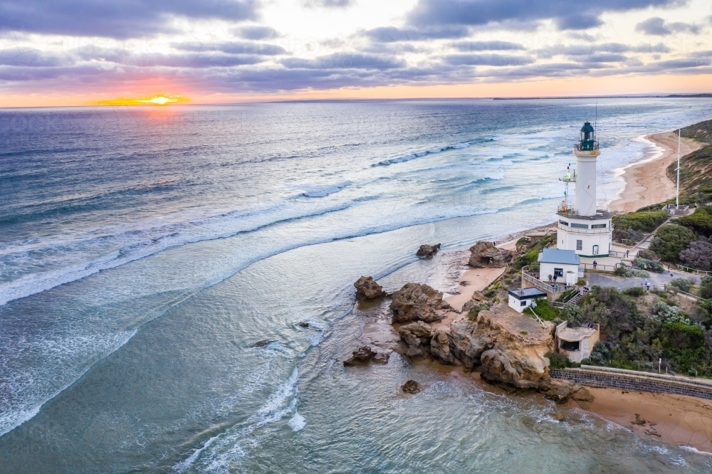 Aerial view of the sun setting off shore from a lighthouse on a cliff top - Australian Stock Image