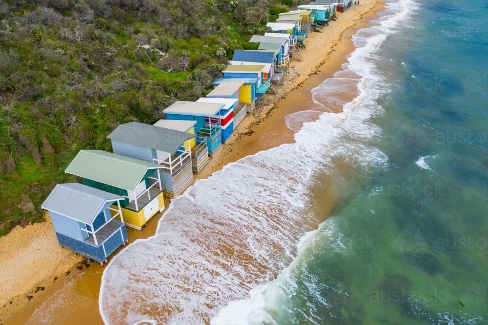 Aerial view of waves crashing on a beach in front of a row of beach huts - Australian Stock Image