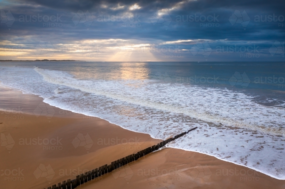 Aerial view of waves washing over a wooden groyne on a sand beach at sunrise - Australian Stock Image
