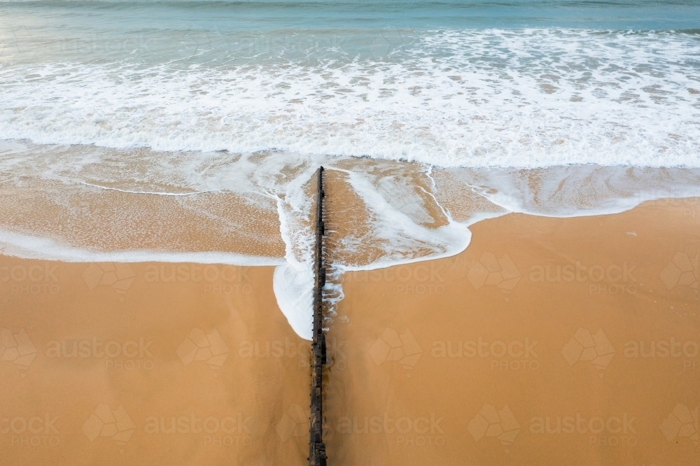 Aerial view of waves washing over a wooden groyne on a sand beach - Australian Stock Image
