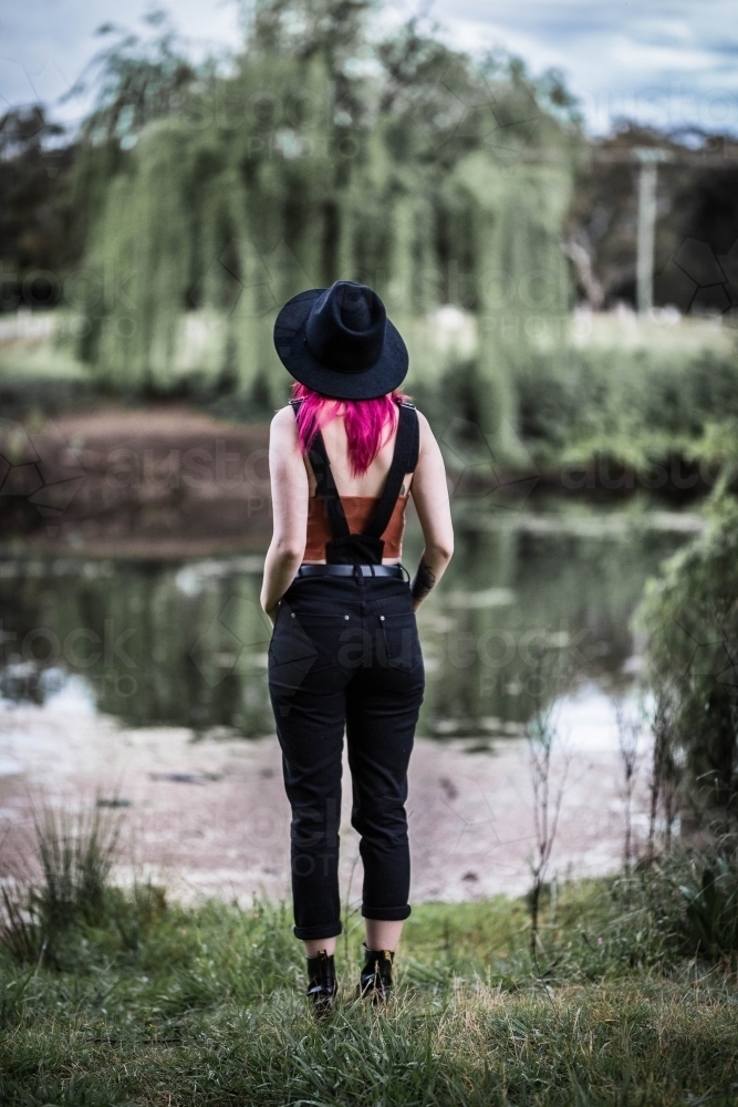 Alternative woman with pink hair and hat in overalls standing looking over river water - Australian Stock Image