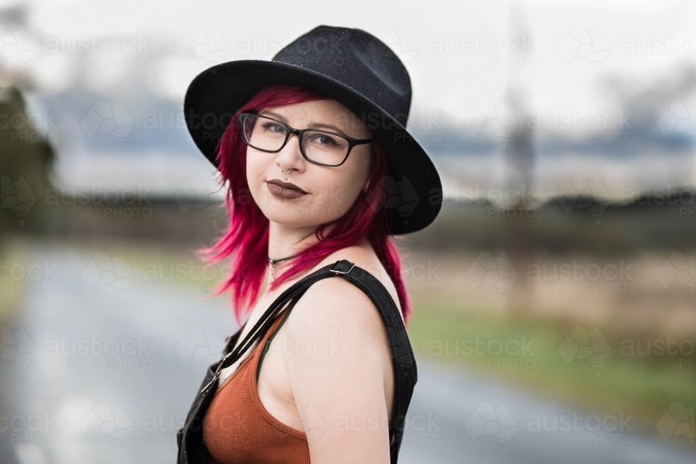 Alternative young woman with glasses and pink hair wearing hat looking over shoulder - Australian Stock Image