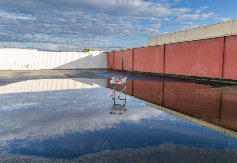 An abandoned shopping trolley in a puddle reflecting the sky and a long  fence - Australian Stock Image
