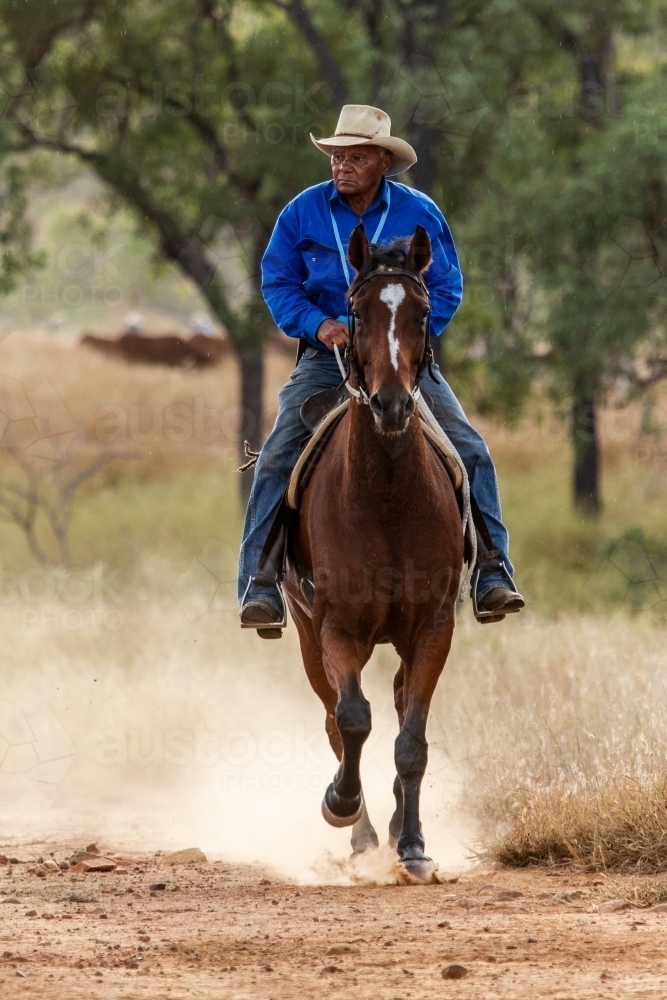An aboriginal stockman cantering on horse kicking up dust - Australian Stock Image