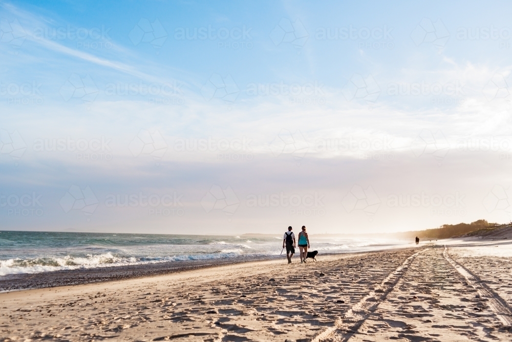 An adult couple and their black dog walk along the beach at sunset, along side tyre tracks - Australian Stock Image