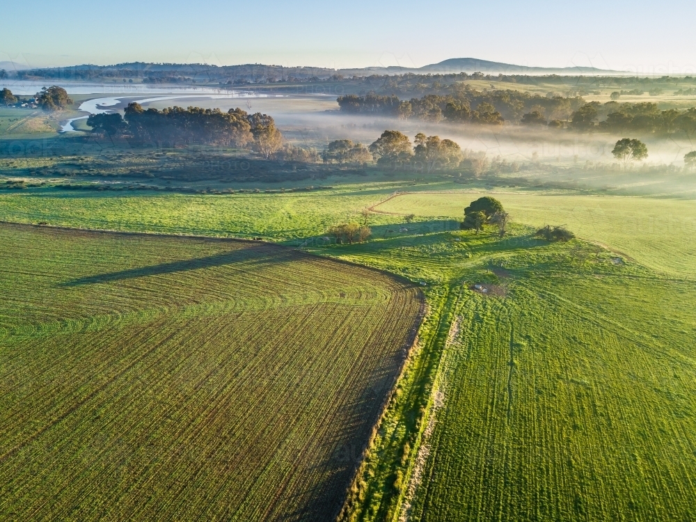 An aerial view of fog over a river running through newly planted green crops - Australian Stock Image