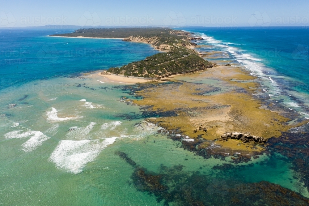 An aerial view of waves crashing onto a rocky platform at the end a a long coastal peninsula - Australian Stock Image