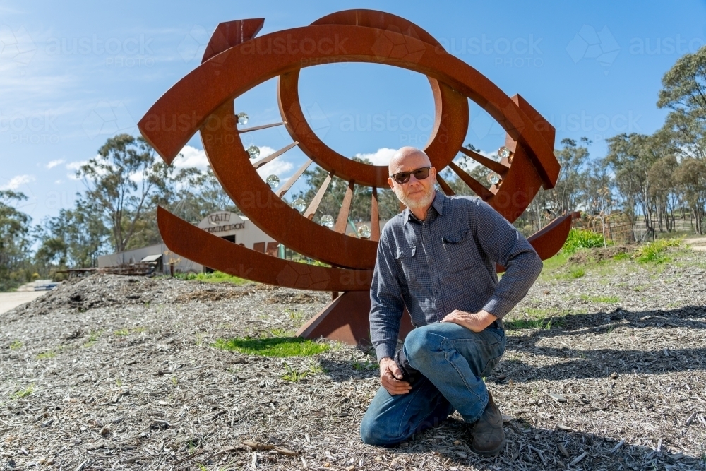 An artist kneeling proudly in front of a large metal sculpture - Australian Stock Image