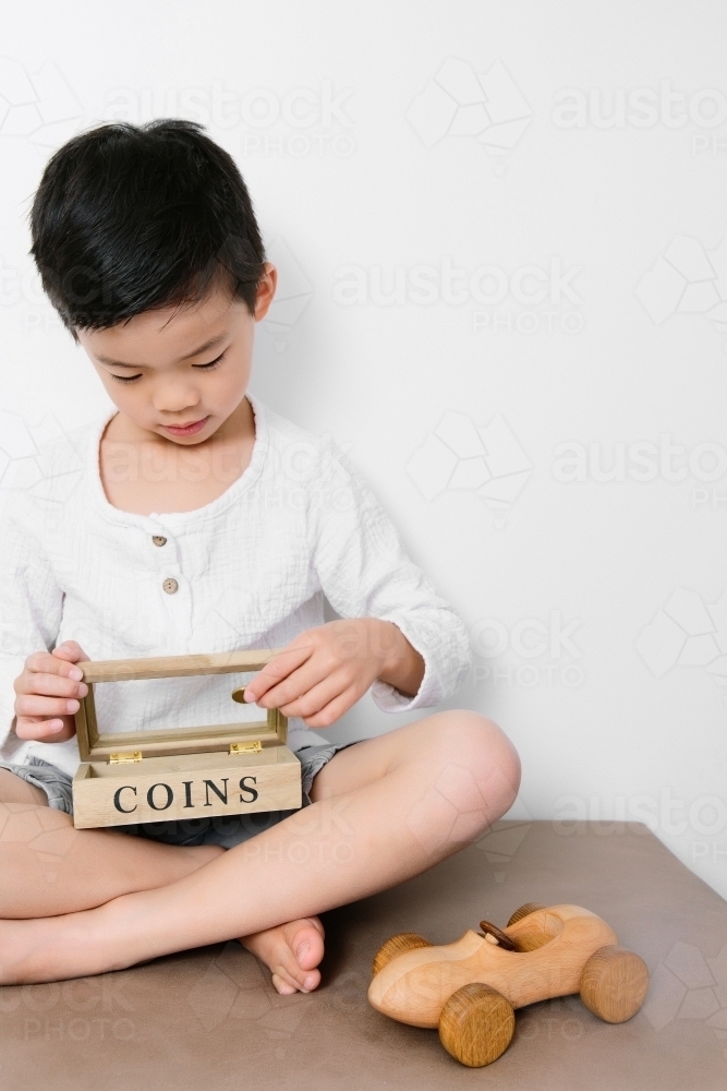 An asian child saving his coins in his money box with a wooden car toy beside him - Australian Stock Image