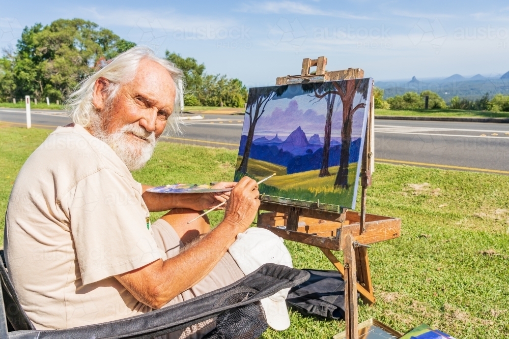 An elderly artist sitting on the roadside painting a canvas on an easel - Australian Stock Image