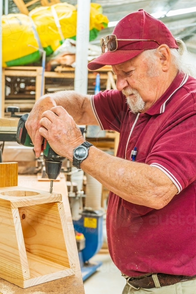 An elderly bearded handyman drilling into woodwork in a Men's shed - Australian Stock Image