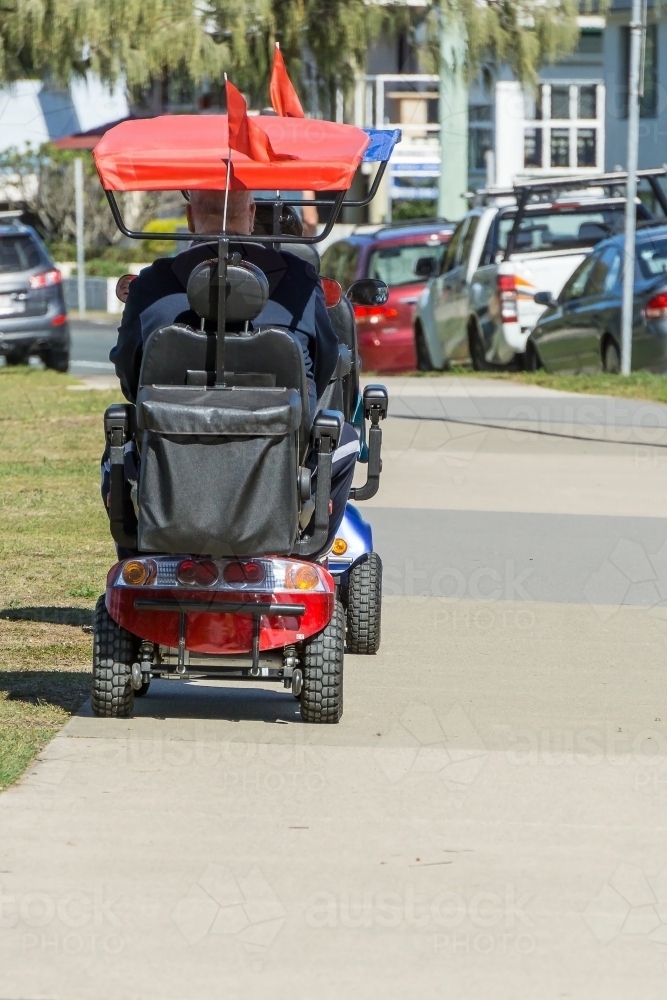 An elderly couple driving mobility scooters along a footpath - Australian Stock Image