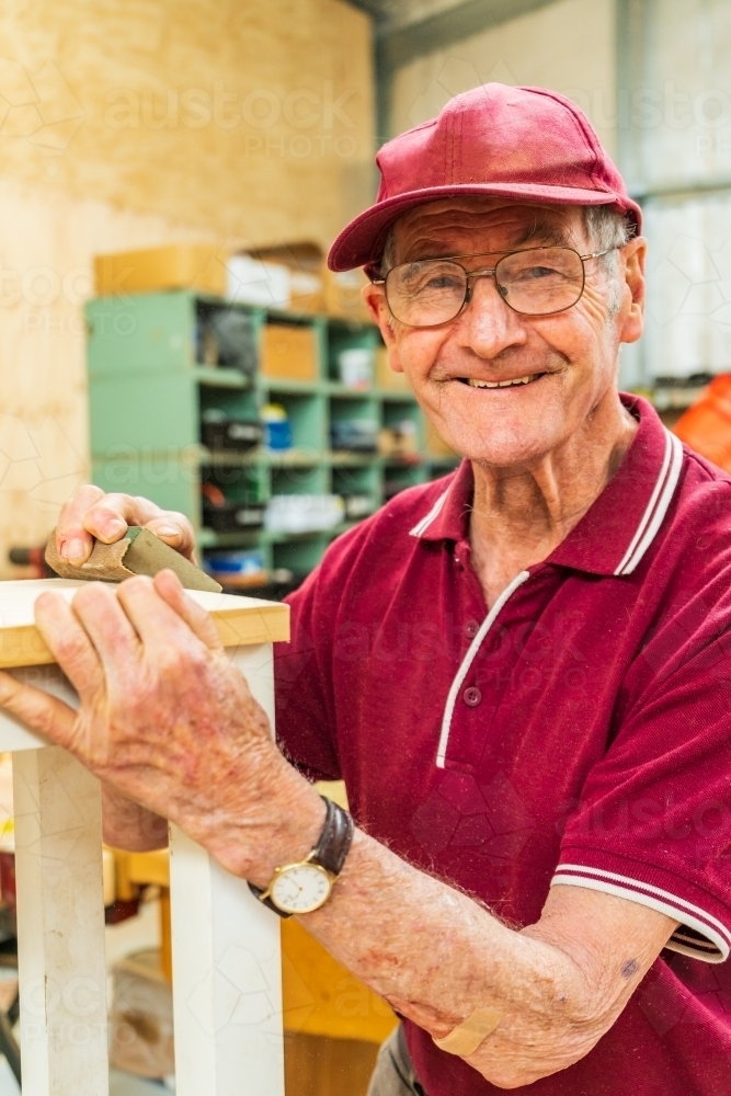 An elderly handyman smiling over the top of a piece of woodwork. - Australian Stock Image