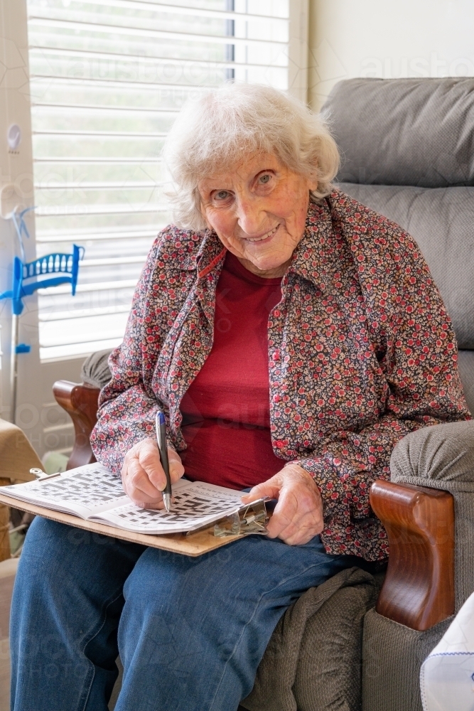 An elderly lady sitting in a chair doing a crossword and smiling - Australian Stock Image