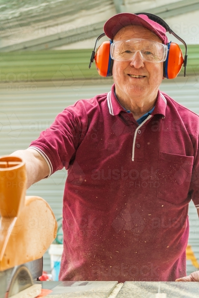 An elderly man smiling behind a bench saw at a Men's shed. - Australian Stock Image