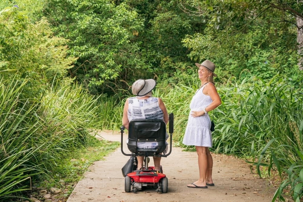 An elderly woman on a disability scooter laughing with her daughter on a path through a park - Australian Stock Image