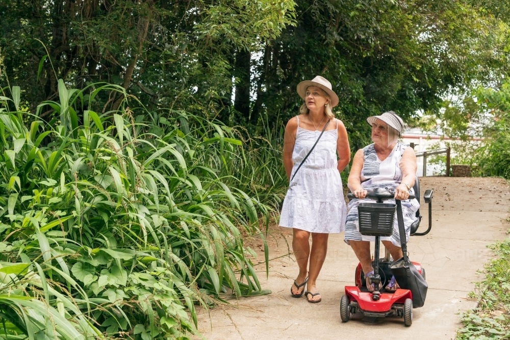 An elderly woman on a disability scooter with her daughter walking down a path in a park - Australian Stock Image