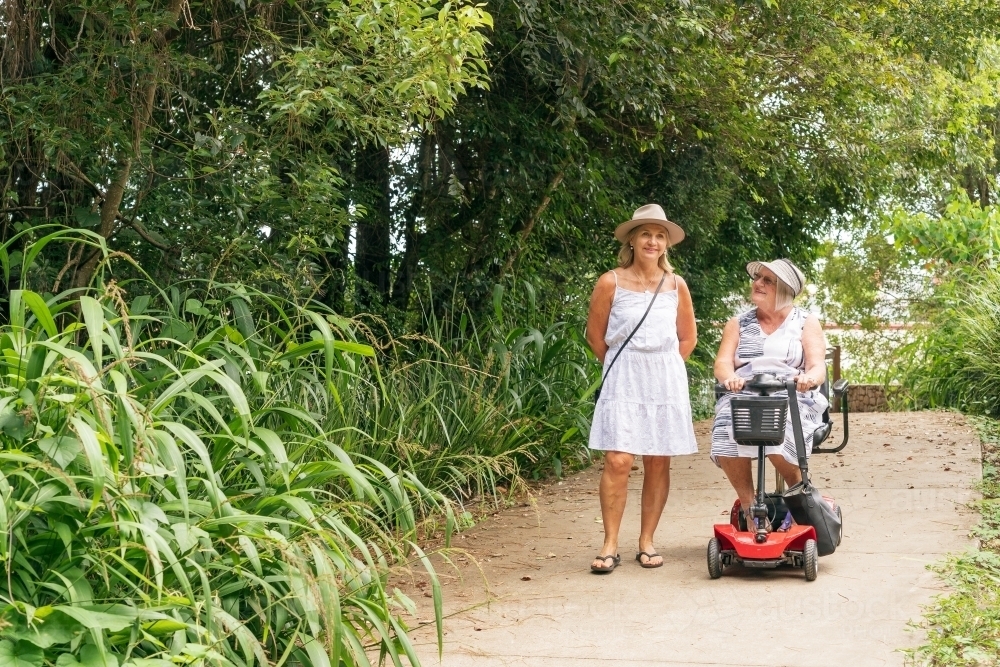 An elderly woman on a disability scooter with her daughter walking down a path in a park - Australian Stock Image