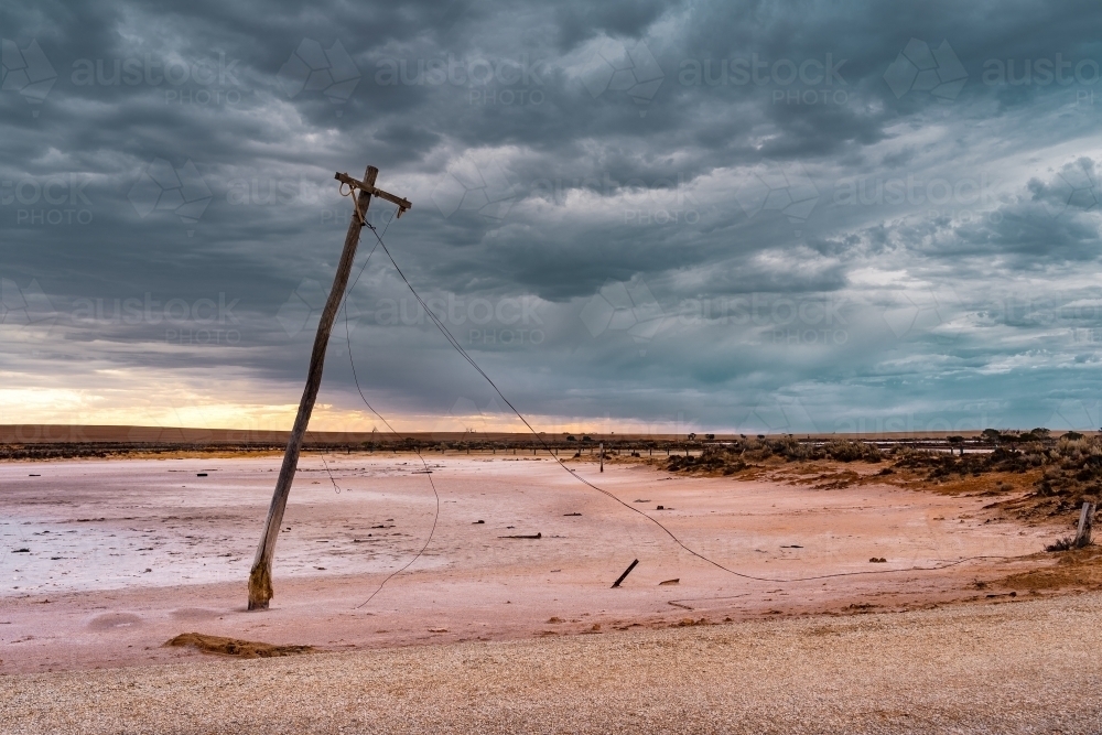 An old power pole leaning over on the edge of a salt lake under a gloomy sky - Australian Stock Image