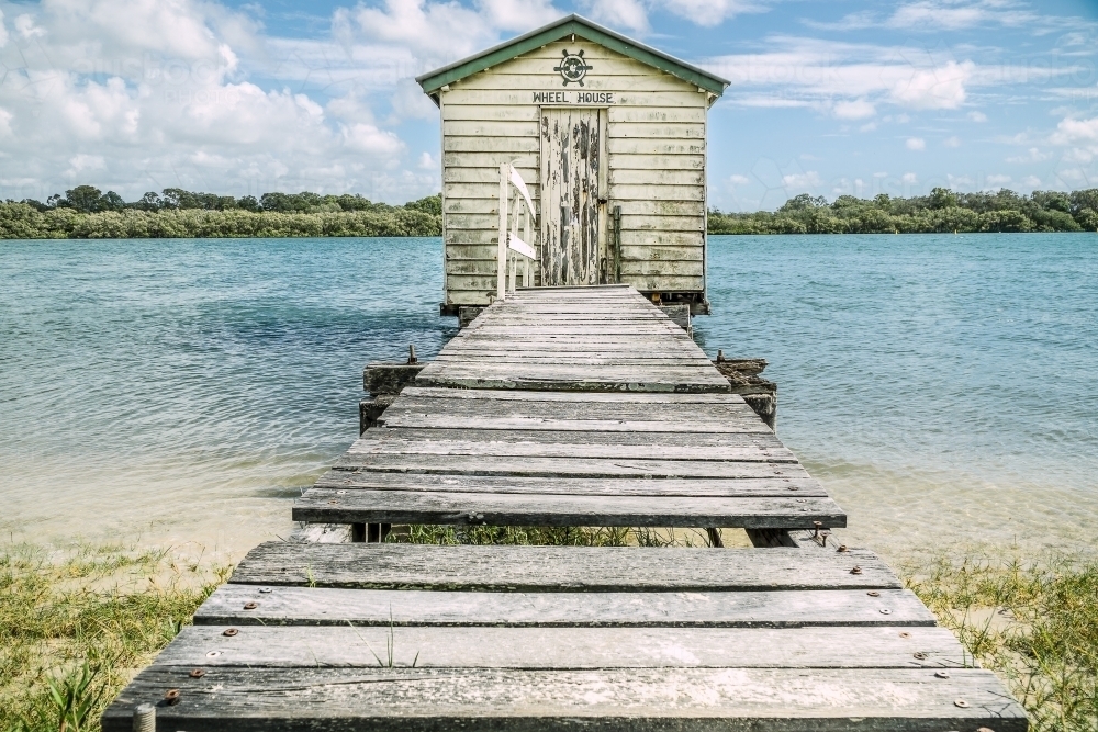 An old wooden jetty leading out to a rustic boatshed over a river - Australian Stock Image