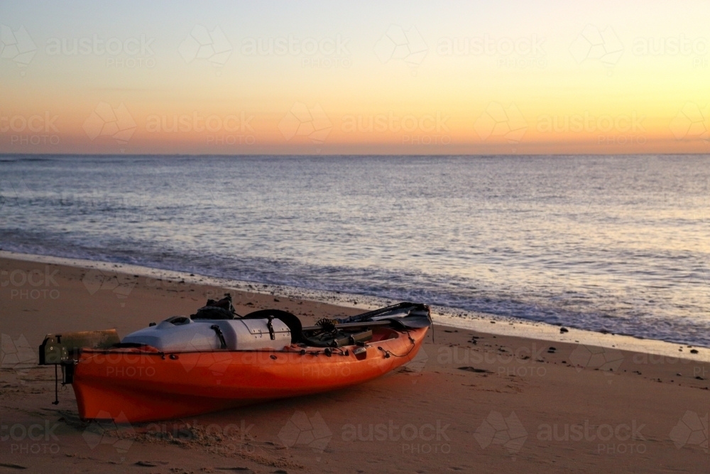 An orange fishing kayak sitting on a beach at sunrise. - Australian Stock Image