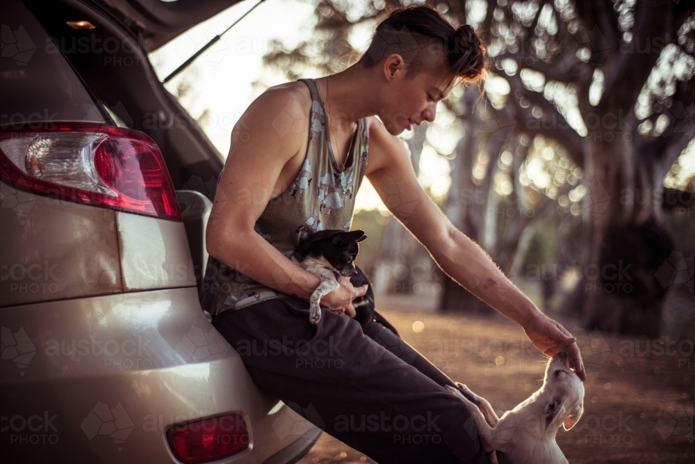 Androgynous woman with two small dogs leans on camping car by river - Australian Stock Image