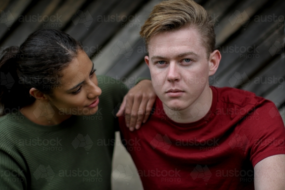 Angry young caucasian male looking at camera while young woman comforts him - Australian Stock Image