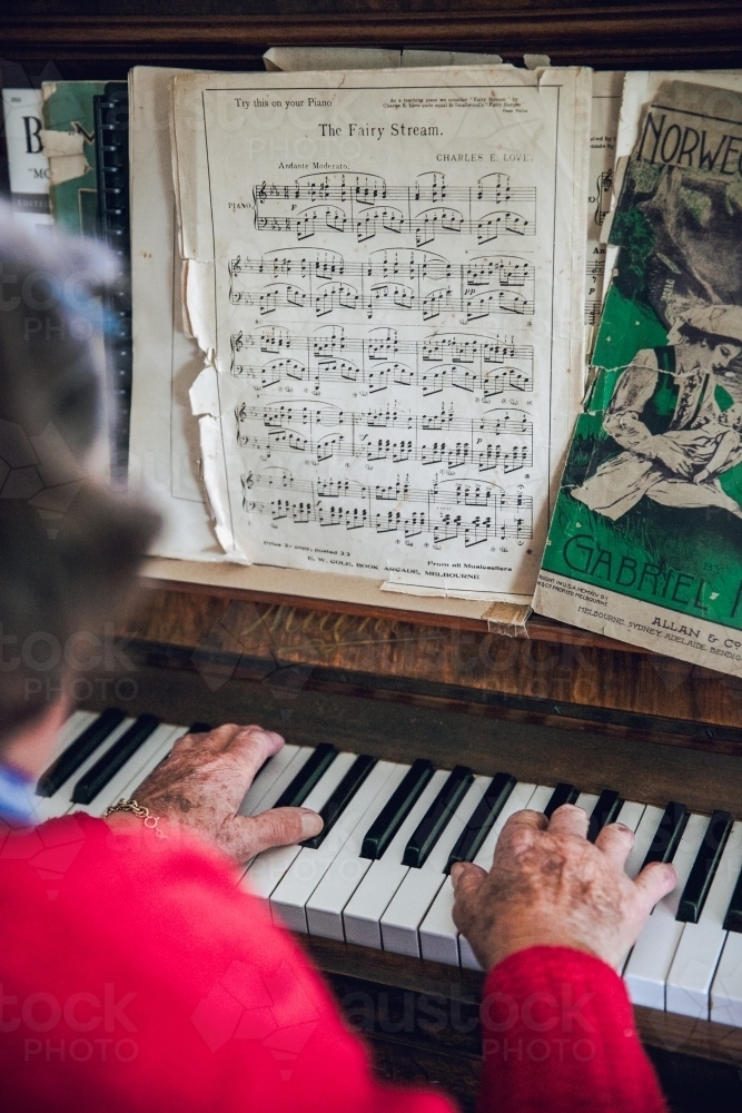 Anonymous old lady playing the piano with sheet music, wearing a red cardigan - Australian Stock Image