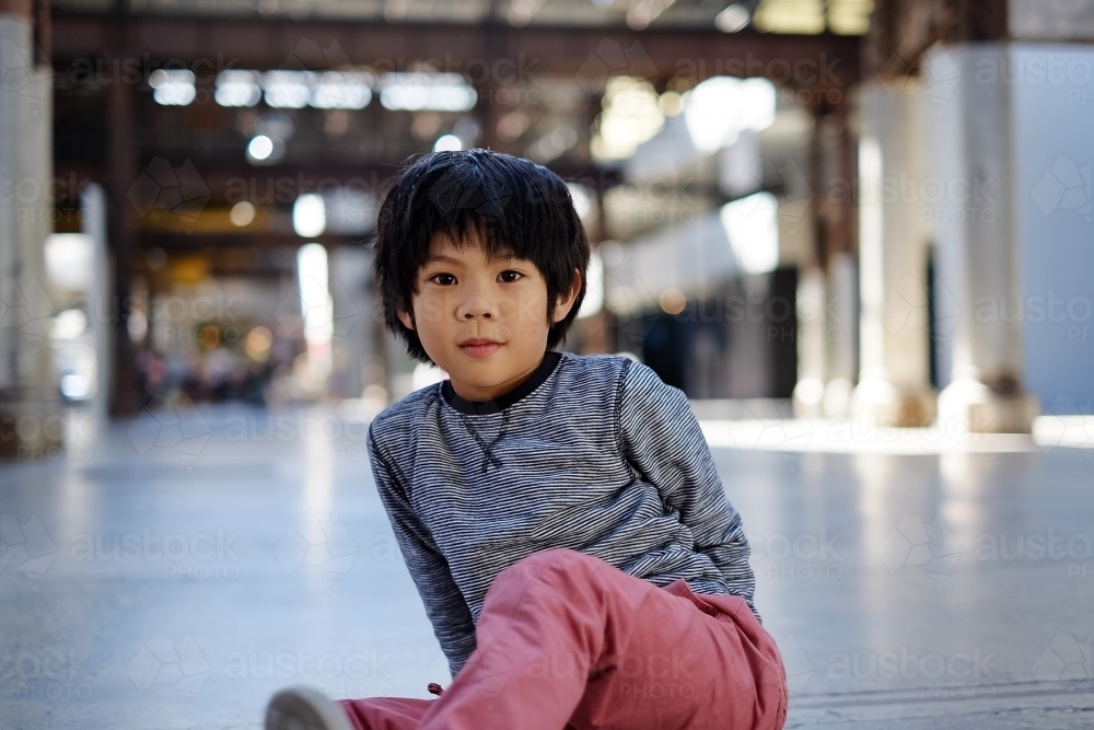 asian boy staring at camera, and sitting on a warehouse floor - Australian Stock Image