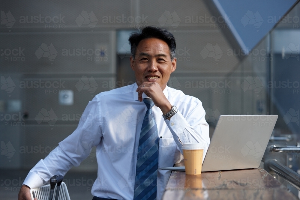 Asian businessman having a coffee whilst using laptop - Australian Stock Image