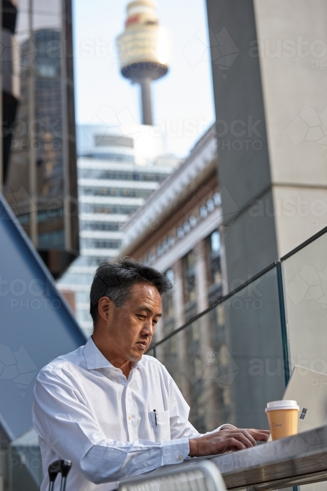 Asian insurance broker working at table outdoors on laptop whilst having a coffee - Australian Stock Image