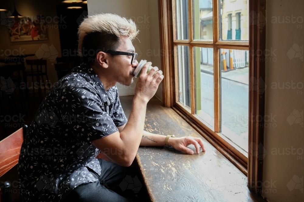 Asian man drinking coffee by the window - Australian Stock Image