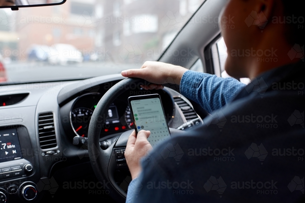 Asian man driving in his car holding mobile phone texting - Australian Stock Image