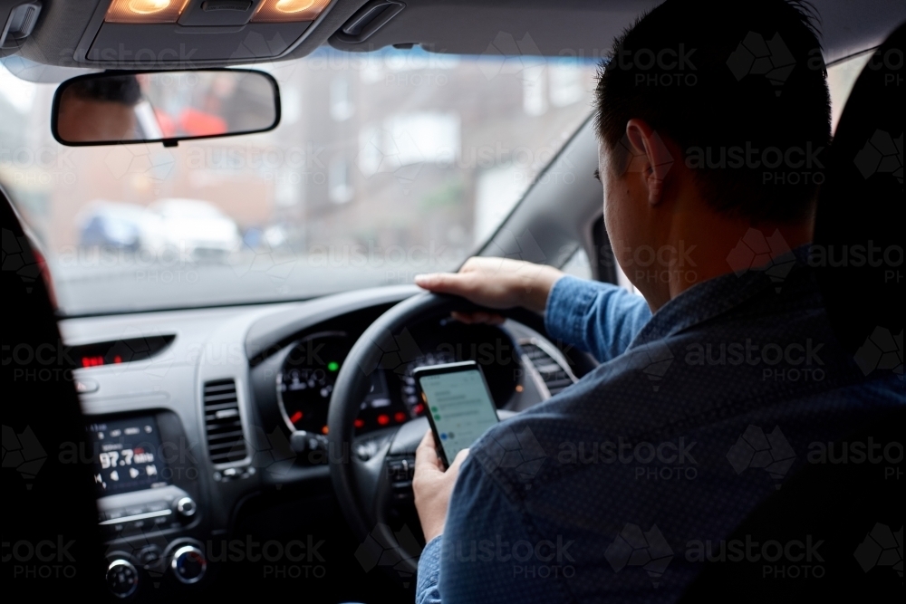 Asian man driving in his car looking down at mobile phone texting - Australian Stock Image
