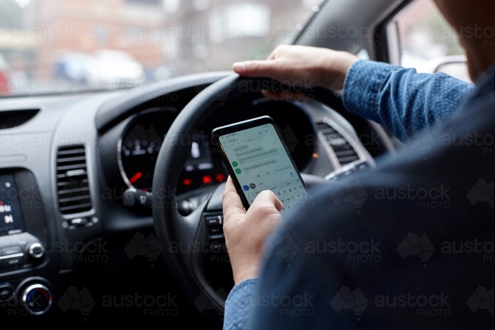 Asian man driving in his car looking down at mobile phone texting - Australian Stock Image