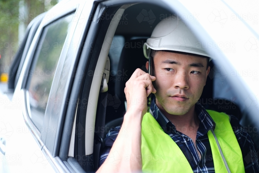 Asian man wearing hard hat in car on phone - Australian Stock Image
