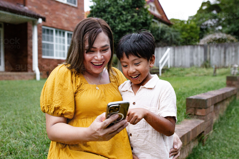 Asian mother and son checking phone while seated on brick wall. - Australian Stock Image