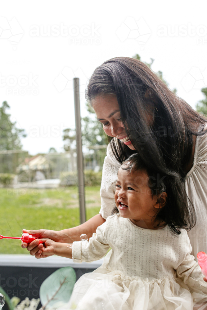 Asian mother with toddler daughter making soap bubbles outdoors - Australian Stock Image