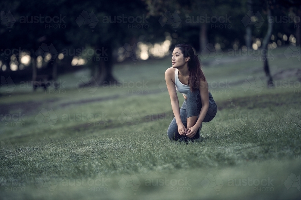 Asian runner tying shoelaces in park during morning exercise - Australian Stock Image