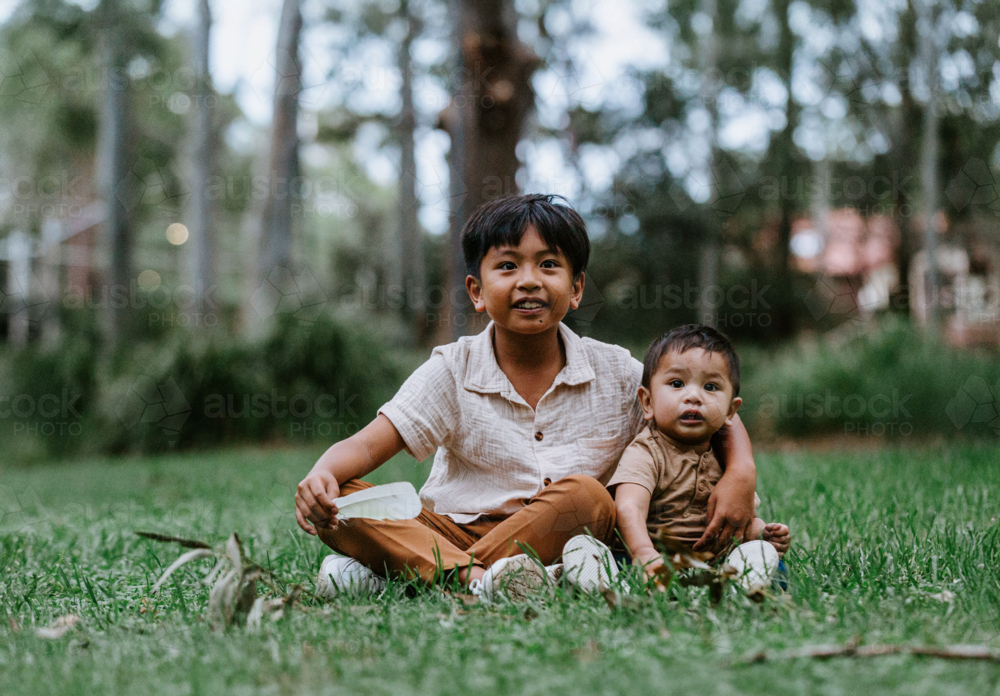 Asian siblings sitting on the grass together at the park - Australian Stock Image