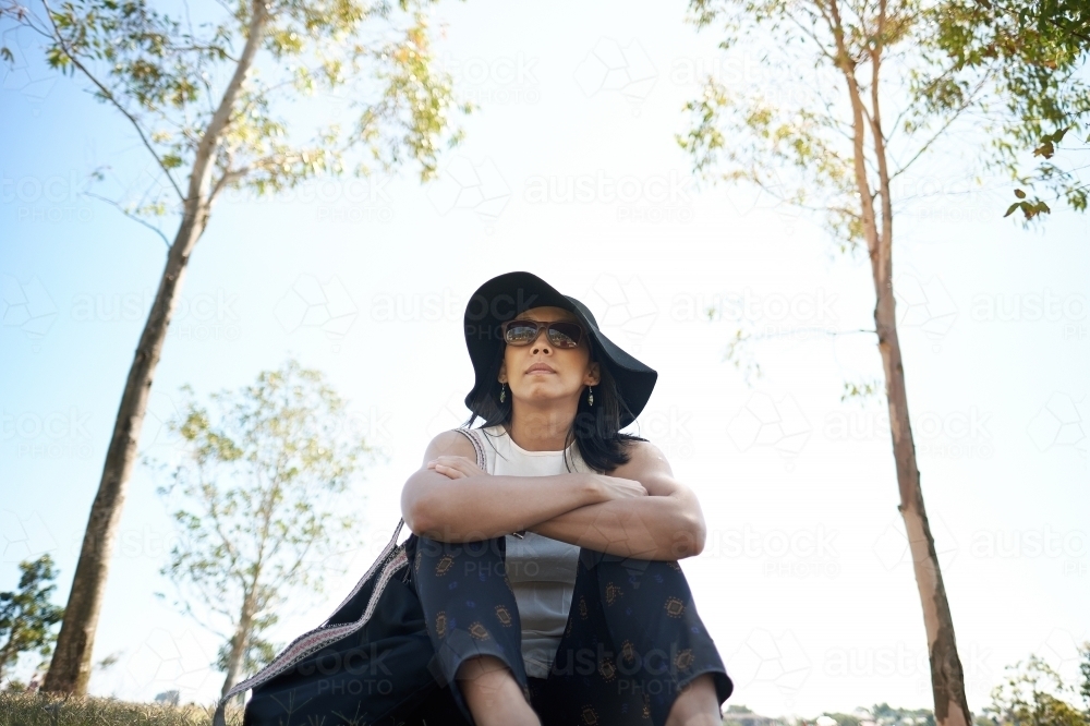 Asian woman sitting at park with sunglasses and hat - Australian Stock Image