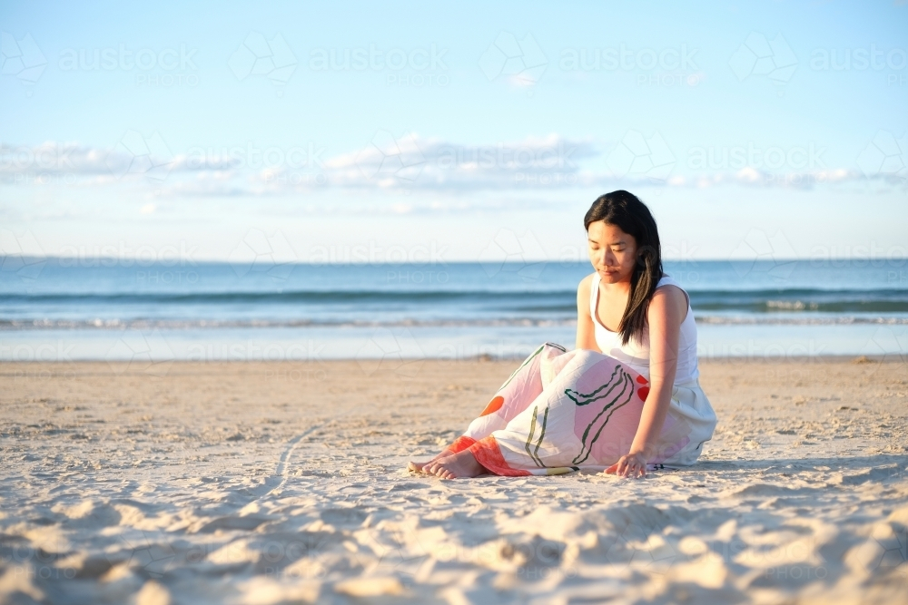 Asian woman sitting on the sand along the beach - Australian Stock Image