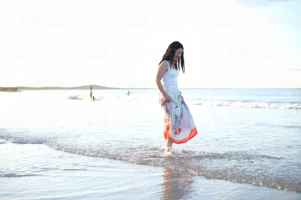 Asian woman walking along the shoreline while holding her skirt up - Australian Stock Image