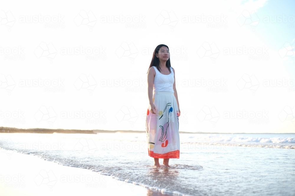 Asian woman wearing a long skirt walking along the beach - Australian Stock Image