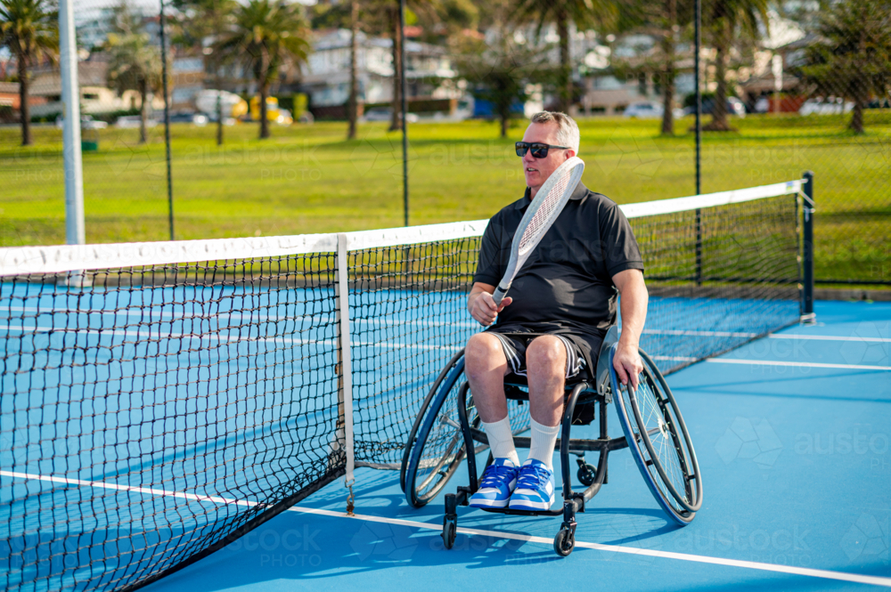 Athlete in wheelchair prepares to serve on a bright day at outdoor tennis facility. - Australian Stock Image