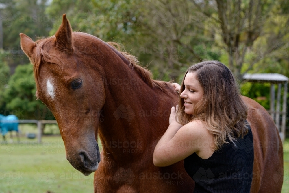Attractive young woman with long brown hair, smiling and rubbing a chestnut horse - Australian Stock Image