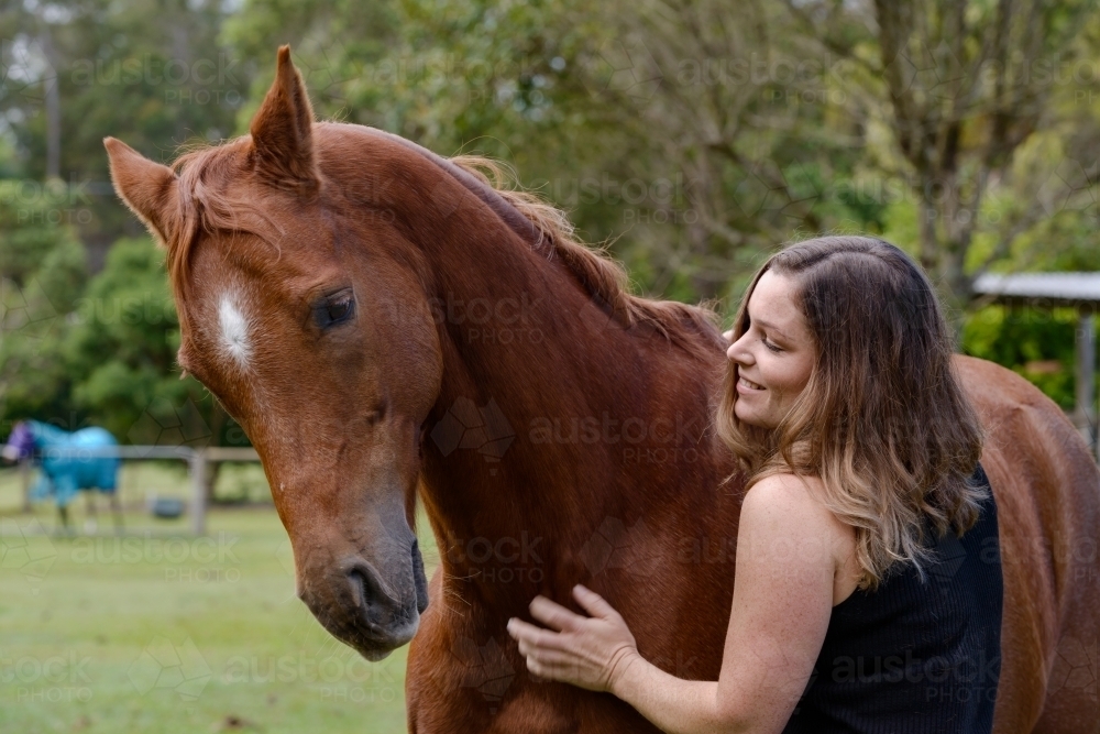 Attractive young woman with long brown hair smiling at a chestnut horse - Australian Stock Image