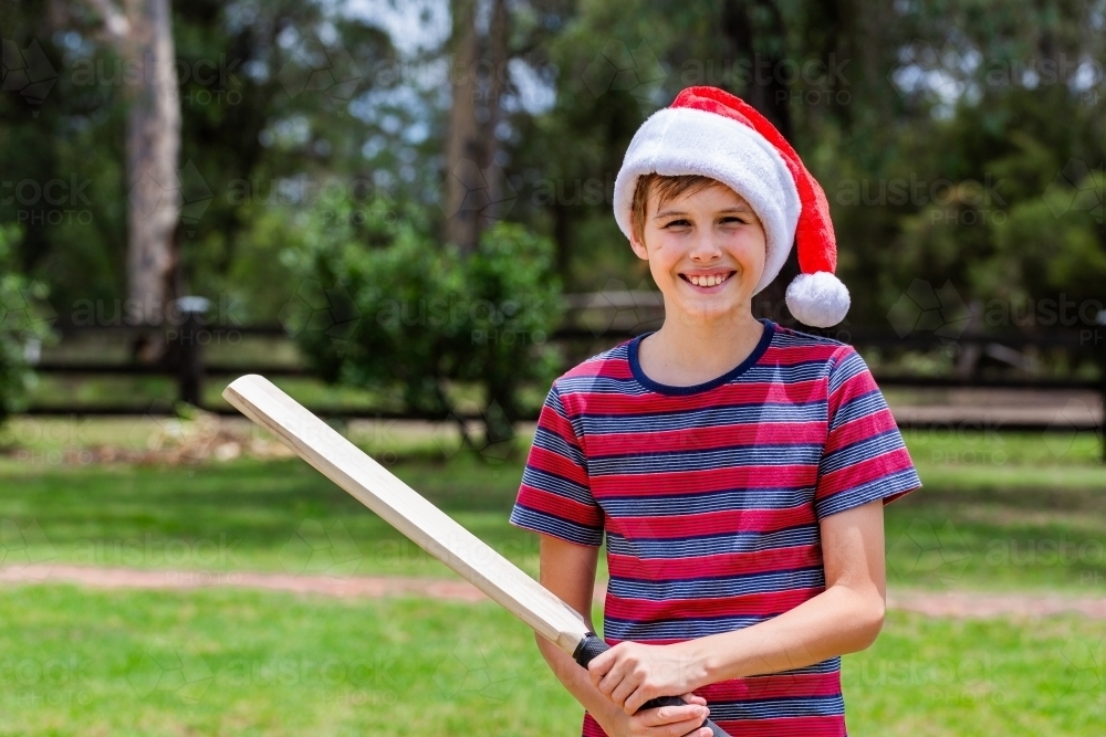 Aussie boy ready to play backyard cricket in front yard at Christmastime wearing santa hat - Australian Stock Image