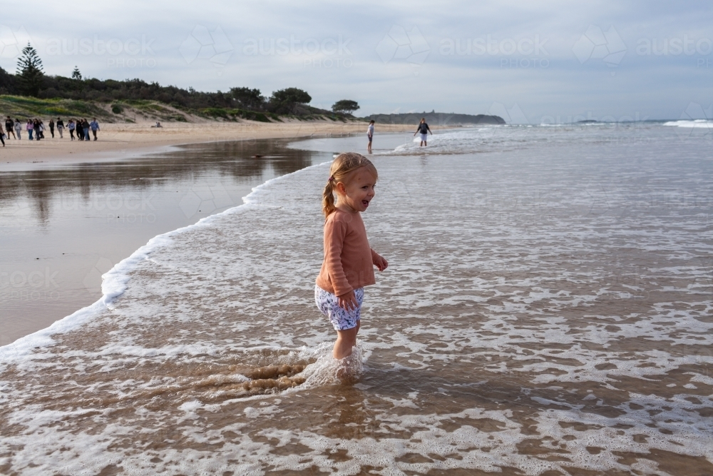 Aussie kid standing in shallow water of beach as waves come in in winter - Australian Stock Image