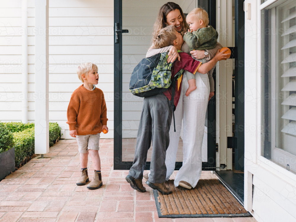 Aussie mum carrying toddler boy while hugging and kissing older son leaving for school at the porch - Australian Stock Image